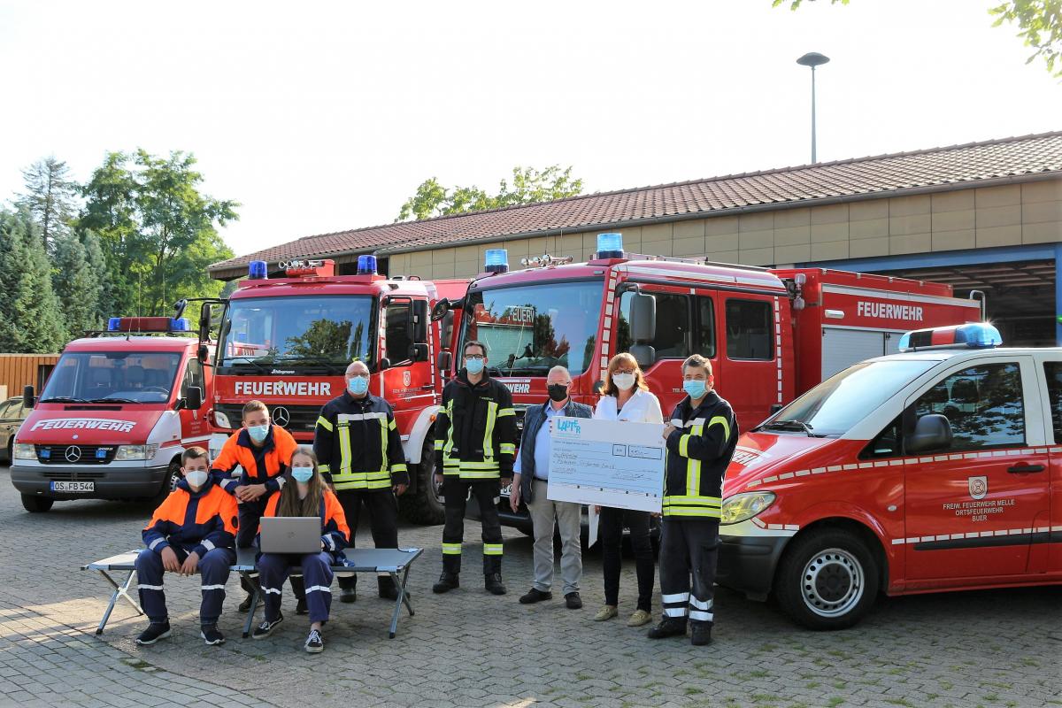 Eine Personengruppe mit einem symbolischen Scheck steht vor vier Feuerwehrfahrzeugen