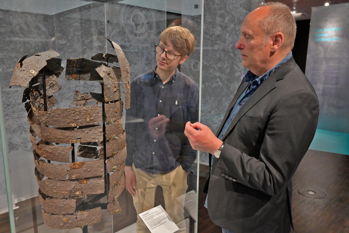 Zwei Personen stehen neben einer Museumsvitrine und schauen auf einen römischen Schienenpamzer.