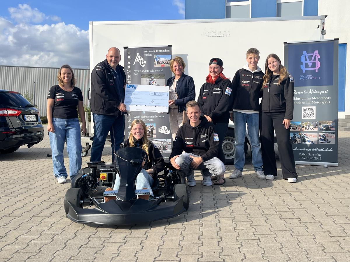 Motorsport Inklusion Eine Personengruppe mit symbolischen Scheck steht um einen E-Kart-Rennwagen herum.