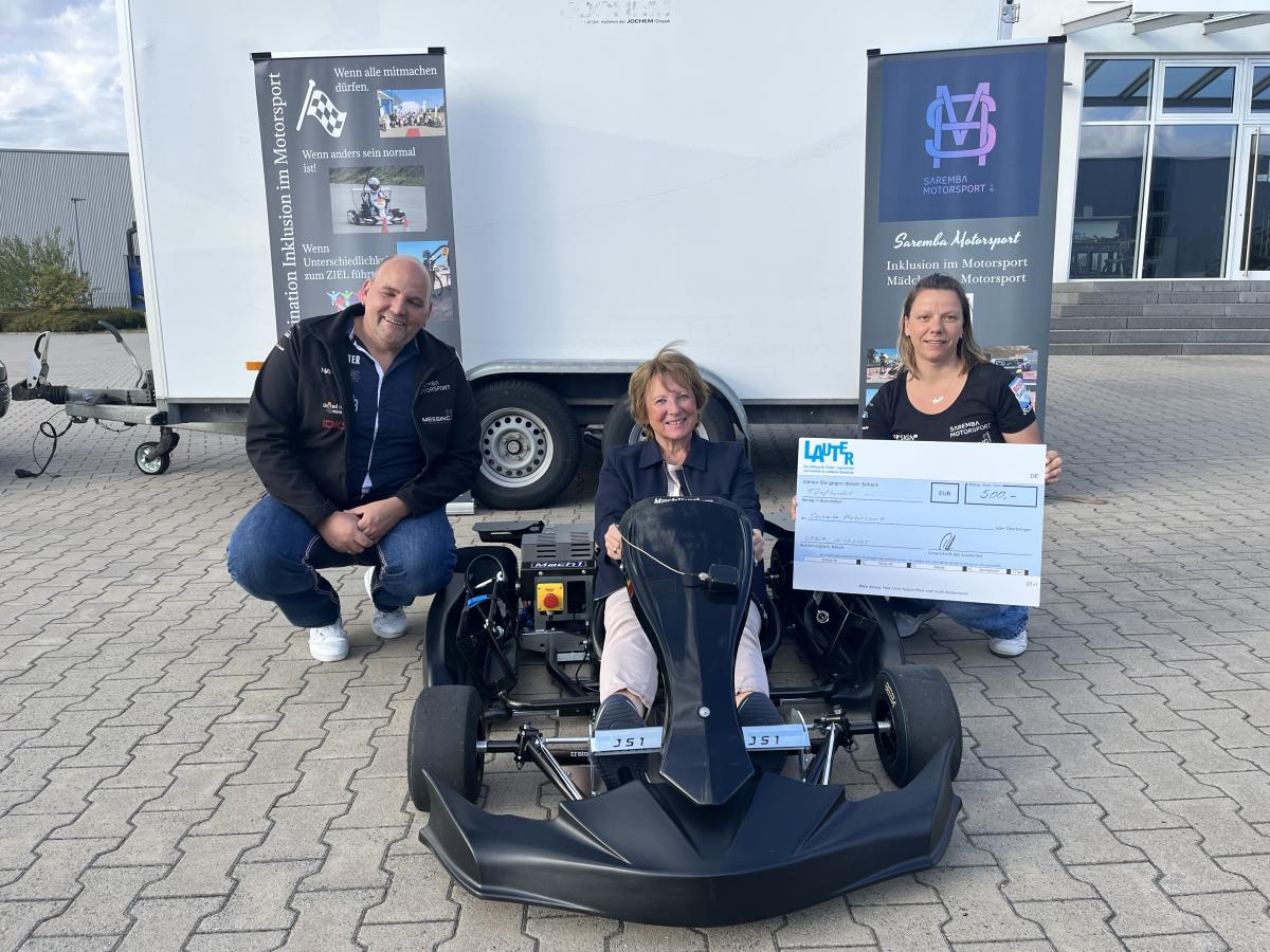 Motorsport Inklusion Eine Personengruppe mit symbolischen Scheck steht um einen E-Kart-Rennwagen herum.