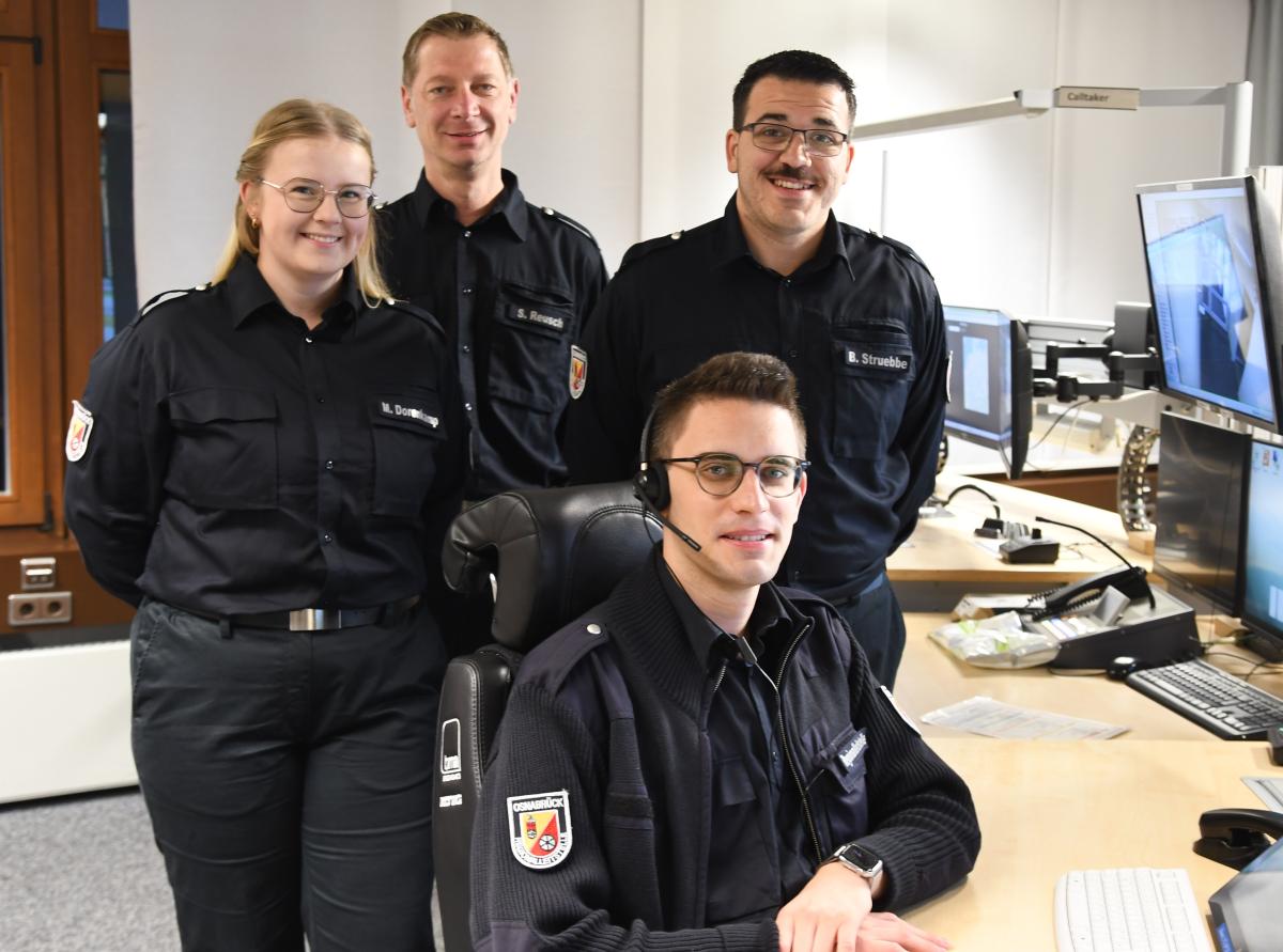 Vier Personen in Uniform stehen und sitzen in einer Rettungsleitstelle.