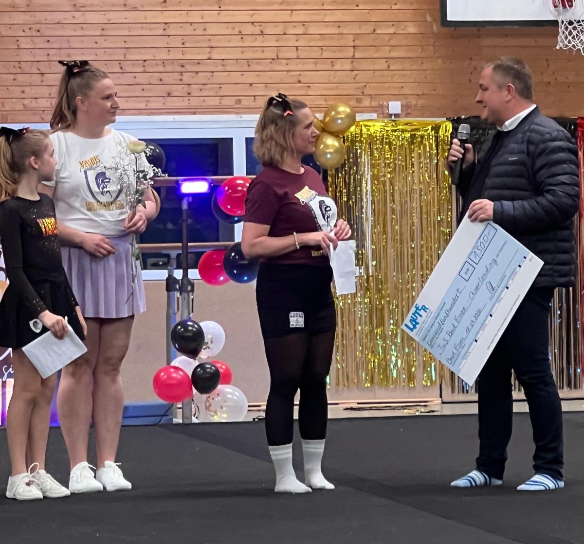 Vier Personen in einer Sporthalle bei einer Spendenscheckübergabe