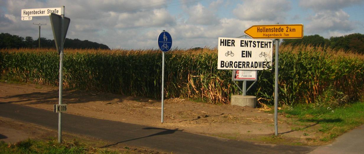 Radweg mit Verkehrsschildern vor einem Maisfeld