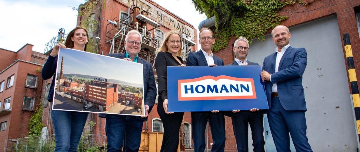 Sechs Personen mit zwei Schildern in den Händen stehen vor einem historischen Fabrikgebäude.