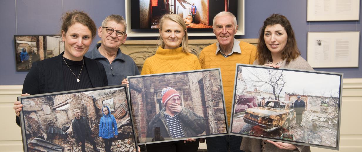 Fünf Personen stehen in einem Raum vor einer Wand und halten großformatige Fotos in den Händen.