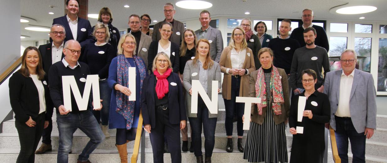 Eine Personengruppe steht in einem Gebäude auf einer Treppe. Einige halten Buchstaben in den Händen, die zusammen das Wort MINT ergeben.