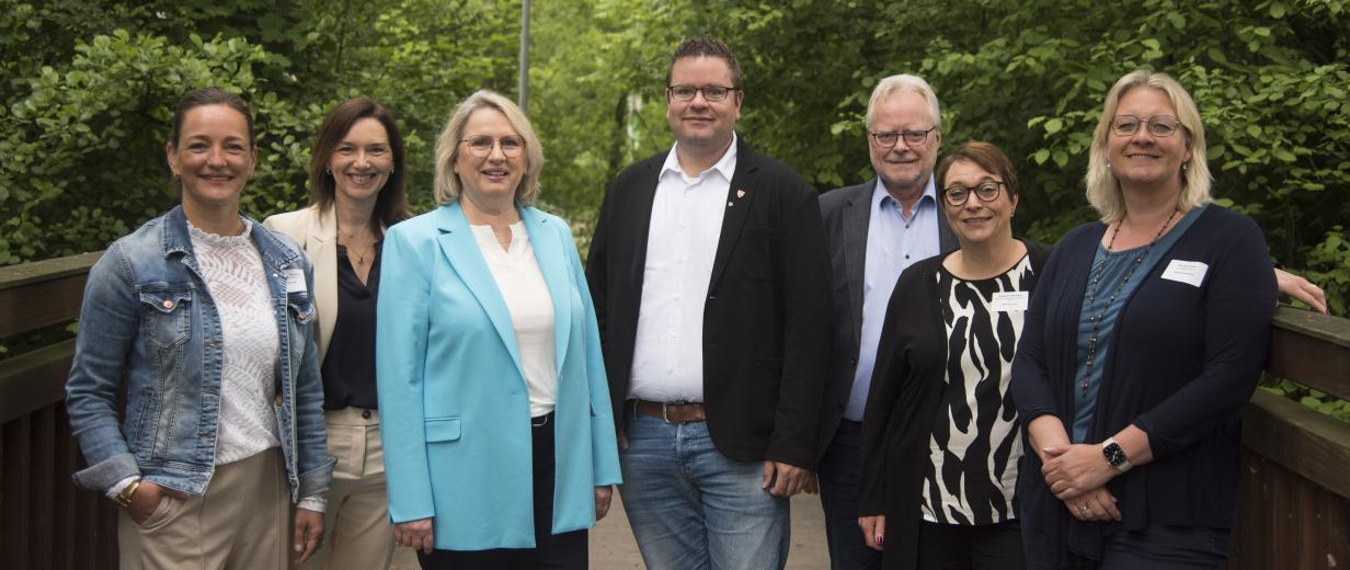 Sieben Personen stehen nebeneinander auf einem Weg mit Bäumen im Hintergrund.