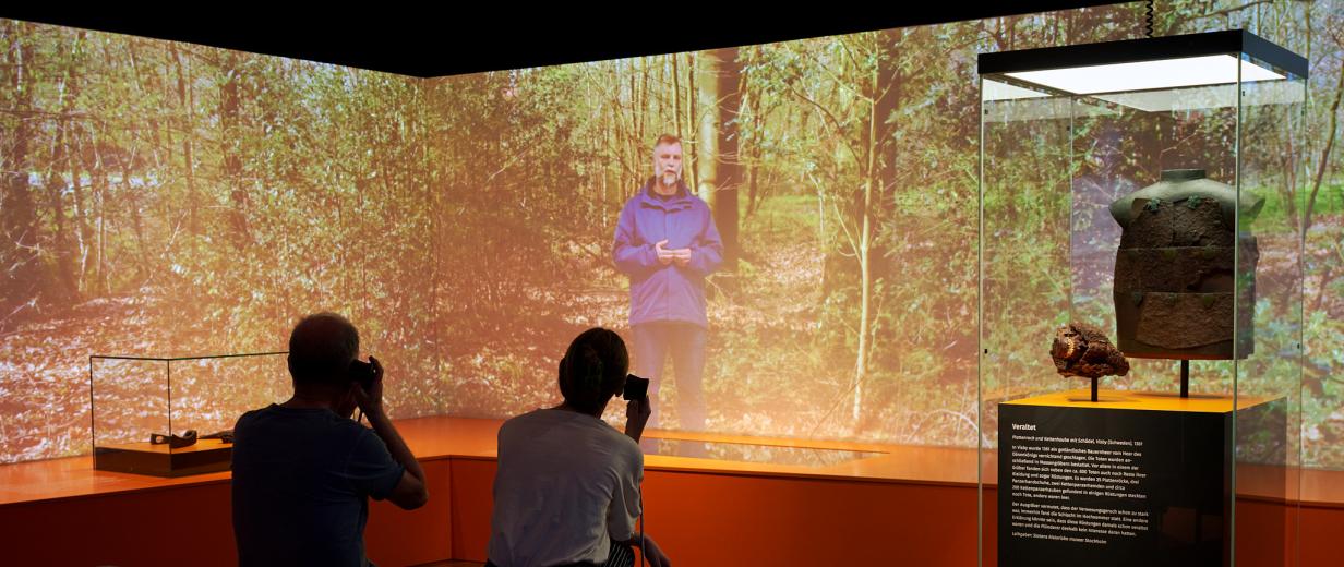 Sonderausstellung 2023 Kalkriese Zwei Personen sitzen vor einer großen Videowand.