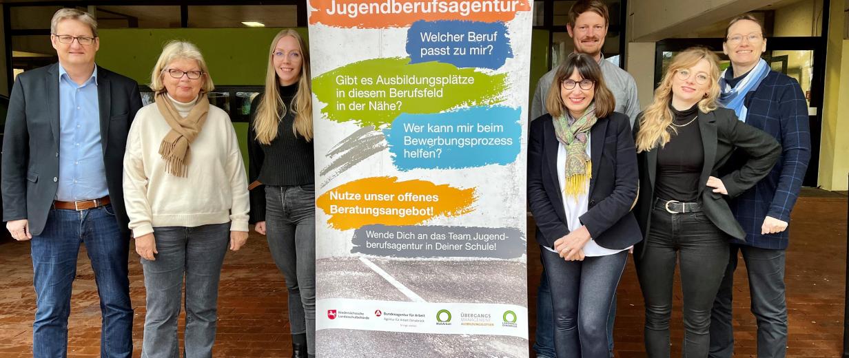 Sieben Personen stehen im Eingangsbereich eines Gebäudes links und rechts neben einem Rollup-Poster.