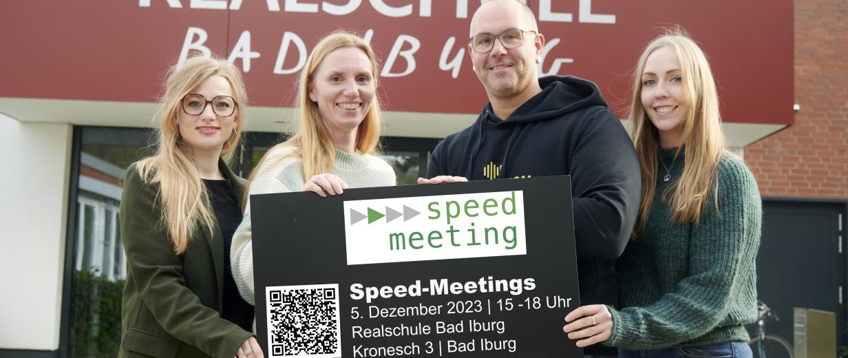 Speed meeting Vier Personen mit einem Schild in den Händen stehen vor einem Gebäudeeingang.