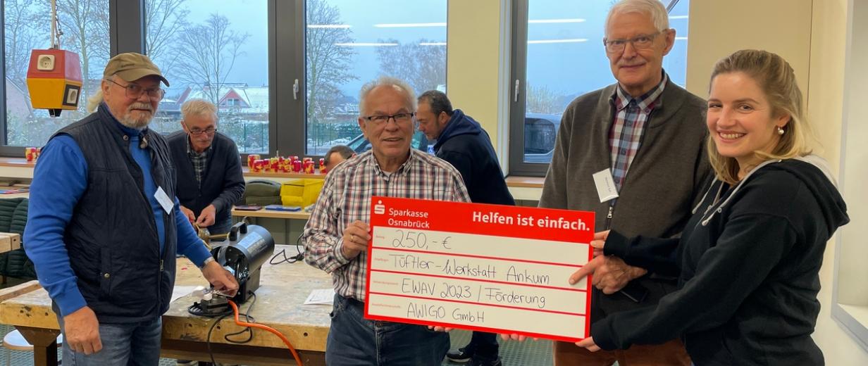 AWIGO Ankum Tüftlerwerkstatt Vier Personen mit einem symbolischen Scheck in den Händen stehen in einer Werkstatt.