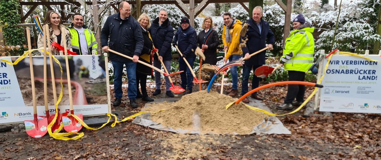 Eine Personengruppe mit Schaufeln steht hinter einem Sandhaufen.