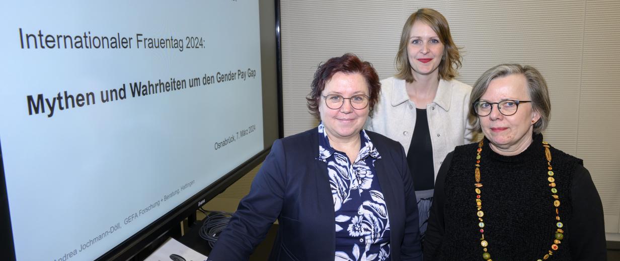 Internationaler Frauentag 2024 Drei Personen stehen neben einem großen Videobildschirm.
