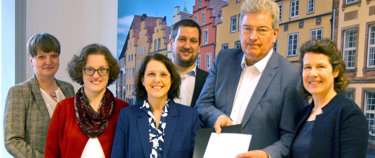 Forschungsprojekt Eingliederungshilfe Hochschule Osnabrück Sechs Personen stehen in einem Raum vor einem großen Foto mit historischen Gebäuden.