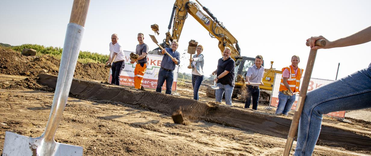 B218, Erweiterung Gerwerbegebiet, oleg Eine Personengruppe mit Spaten in den Händen steht auf einer Baustelle.