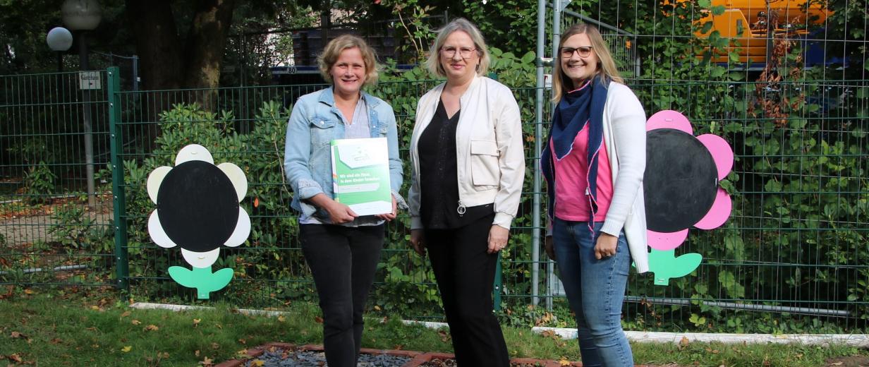 MINT, KiTa, Auszeichnung Drei Personen stehen vor einem geschmückten Zaun.