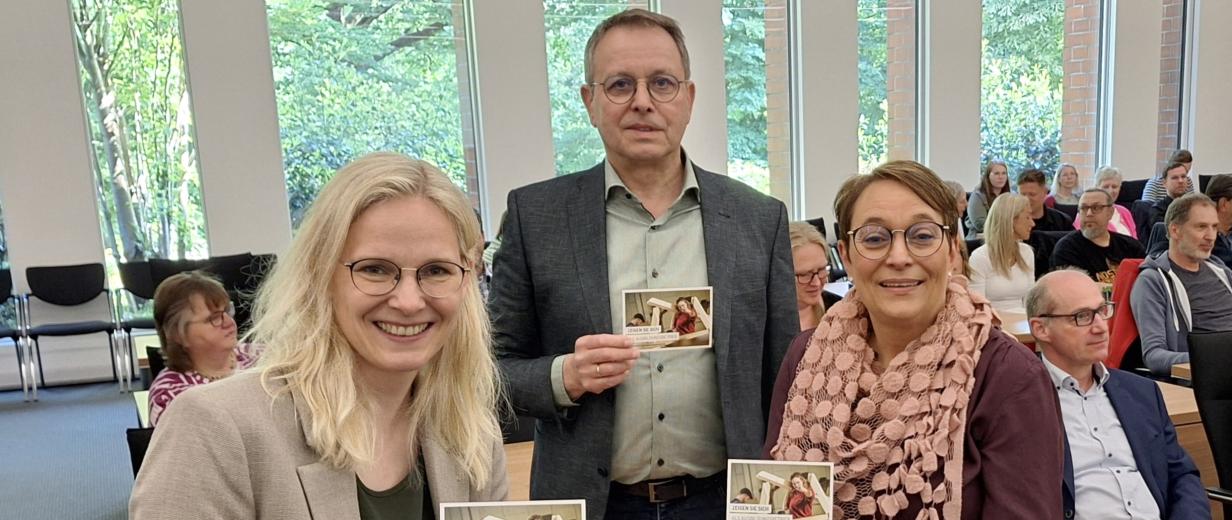 Drei Personen in einem Versammlungssaal halten Flyer in die Kamera.