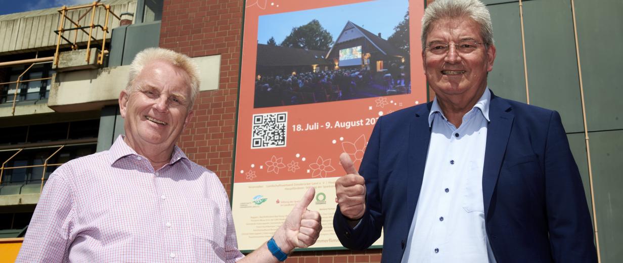 Zwei Personen stehen vor einem großen Plakat an einer Fassade.