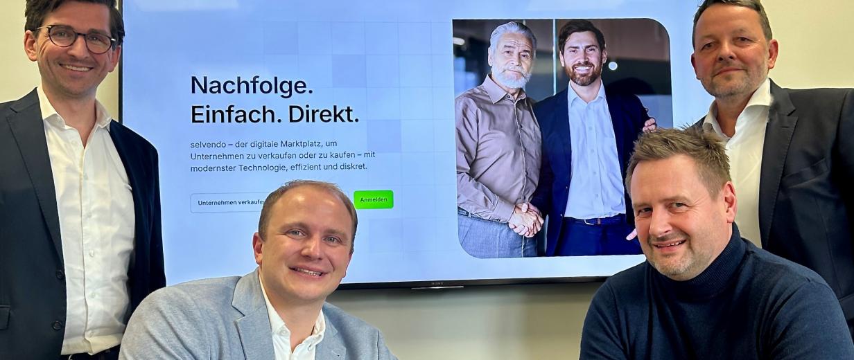 Nachfolge WIGOS selvendo Vier Personen sitzen vor und stehen neben einem großen Bildschirm.