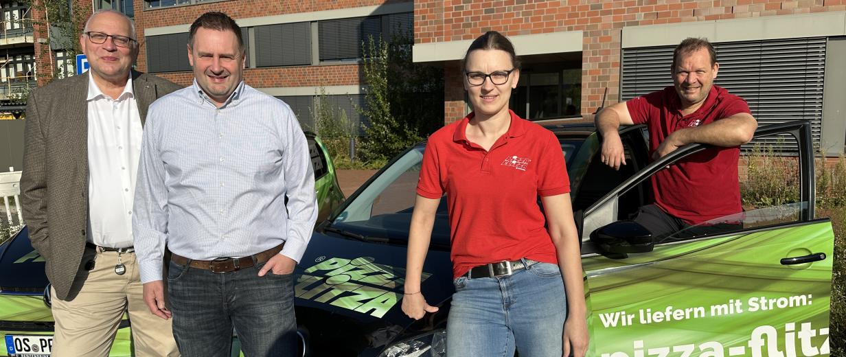 Vier Personen stehen neben dem Auto eines Pizzalieferdienstes.
