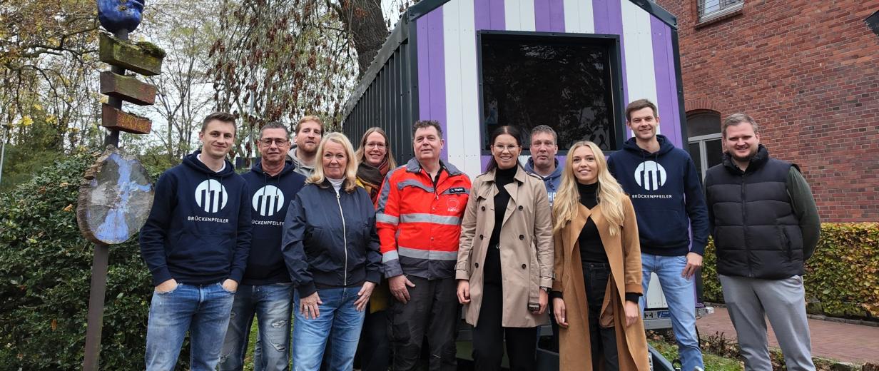 Personengruppe vor einem kleinen Holzhaus