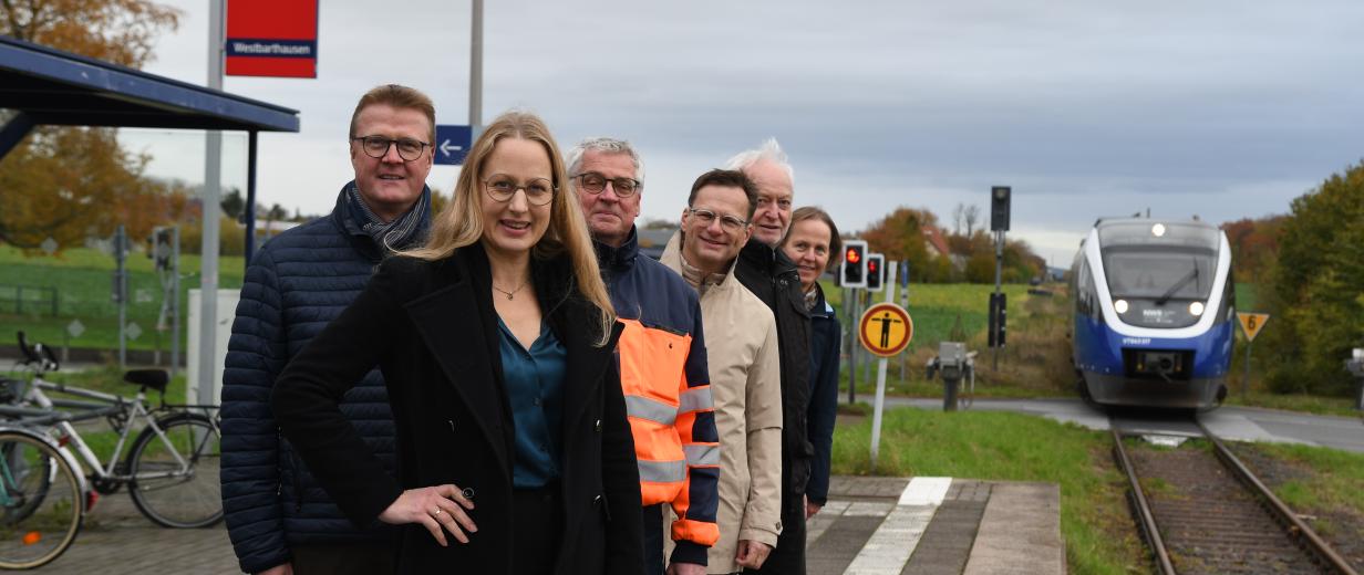 Eine Personengruppe steht auf einem Bahnsteig. Im Hintergrund nähert sich ein Zug.