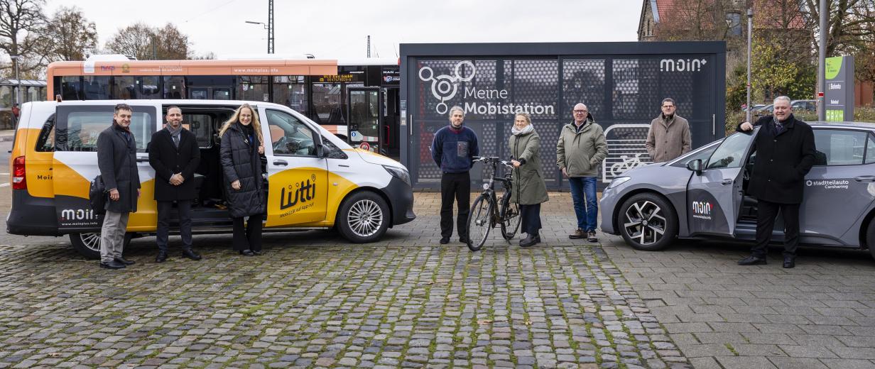 Eine Personengruppe steht vor einem Kleinbus und einem PKW.
