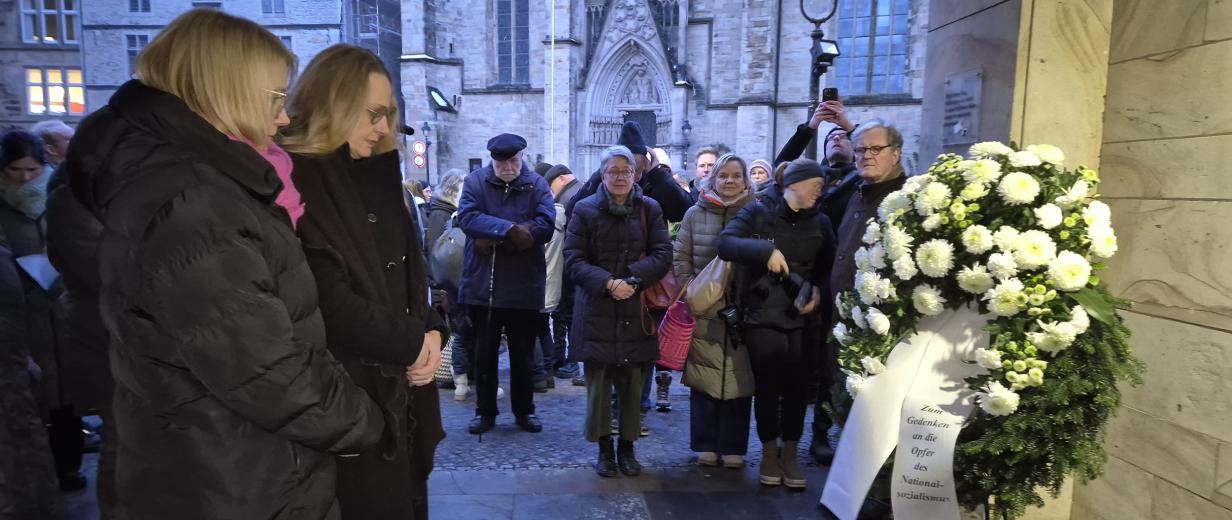 Personen stehen vor einem Gedenkranz.