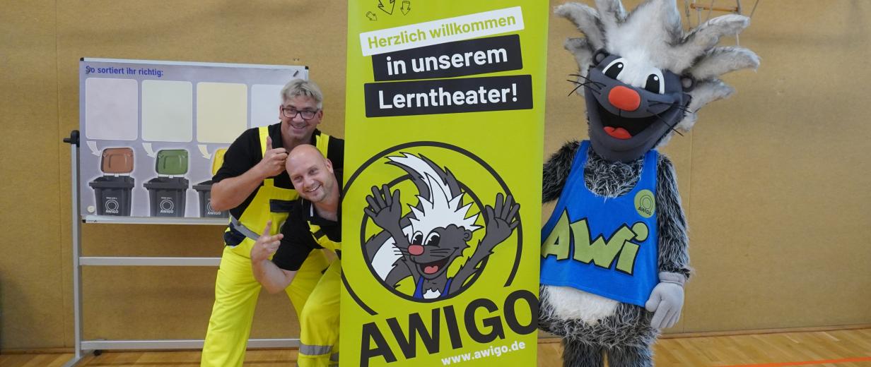 Zwei Personen und ein Maskottchen stehen neben einem Roll Up Poster.