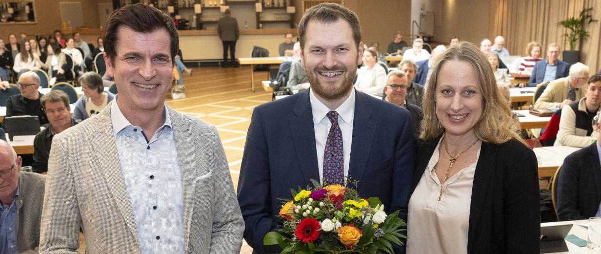 Drei Personen stehen in einem Sitzungssaal.