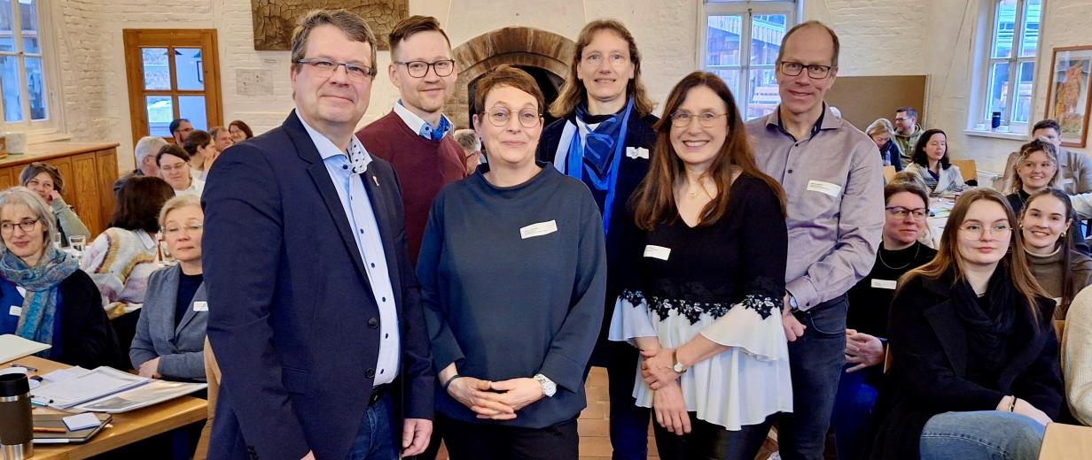 Sechs Personen stehen in einem Vortragsraum zwischen Stuhlreihen