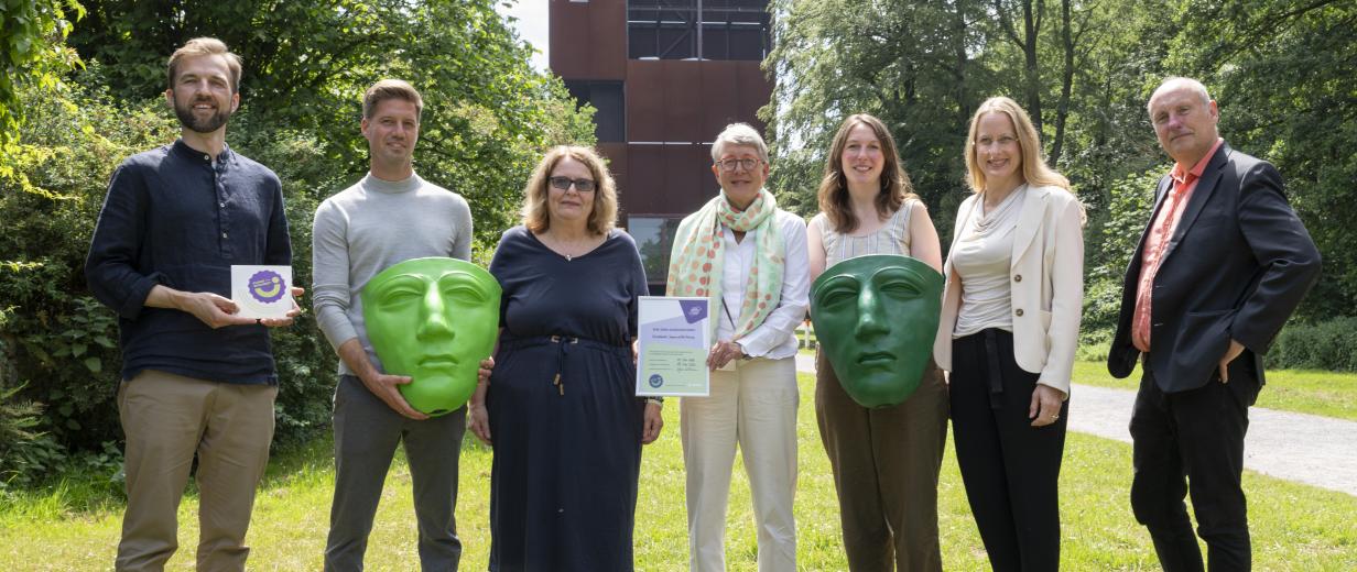 Sieben Personen mit großen römischen Masken stehen auf einer Wiese.