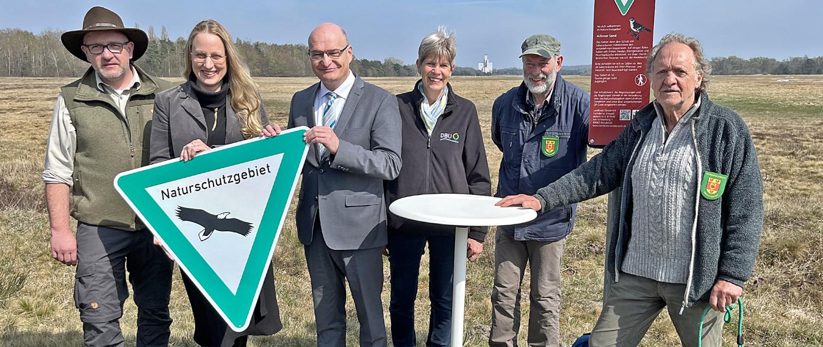 Eine Personengruppe steht am Rande eines Heidegebietes und hält ein Naturschutzschild in die Kamera.