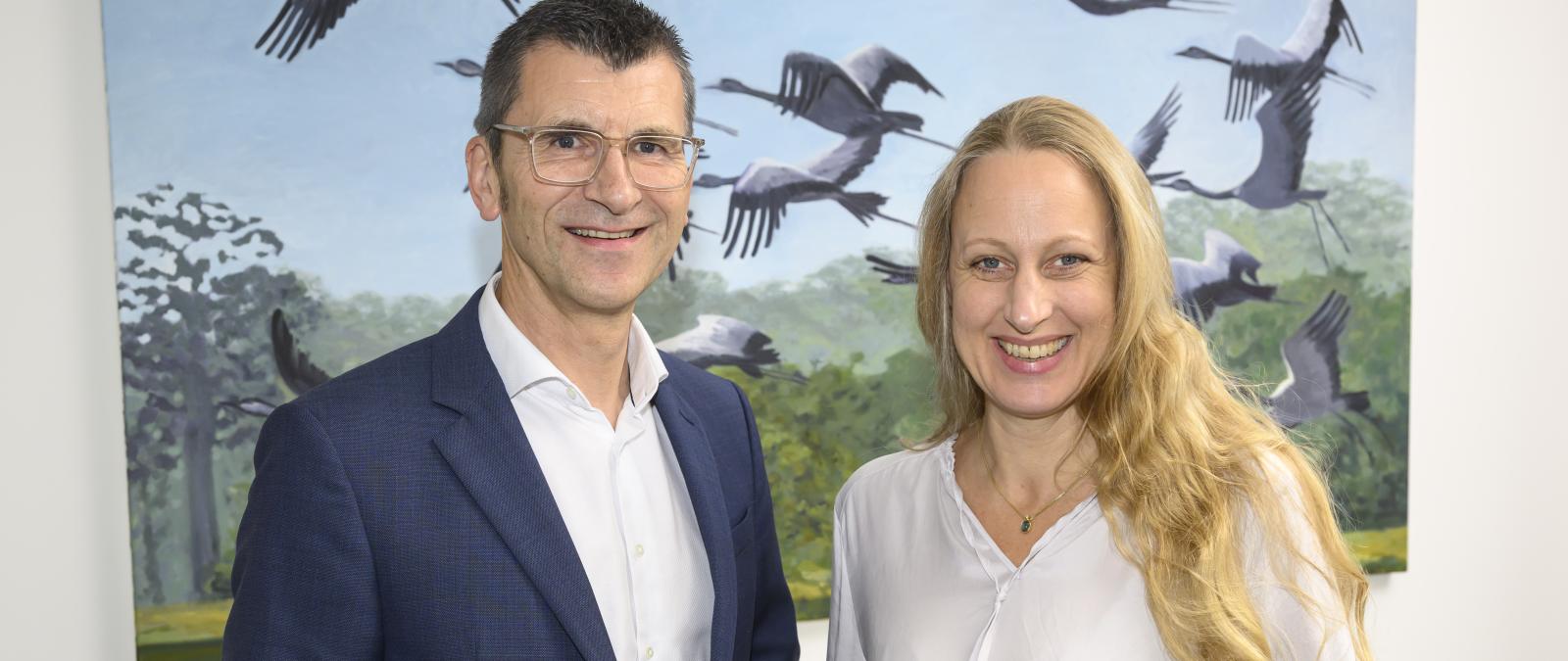 Zwei Personen stehen nebeneinander im Hintergrund ein Bild mit Vögeln.