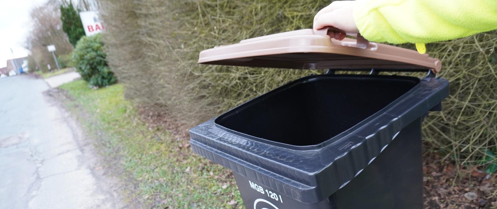 Person hebt den braunen Deckel einer Abfalltonne an.