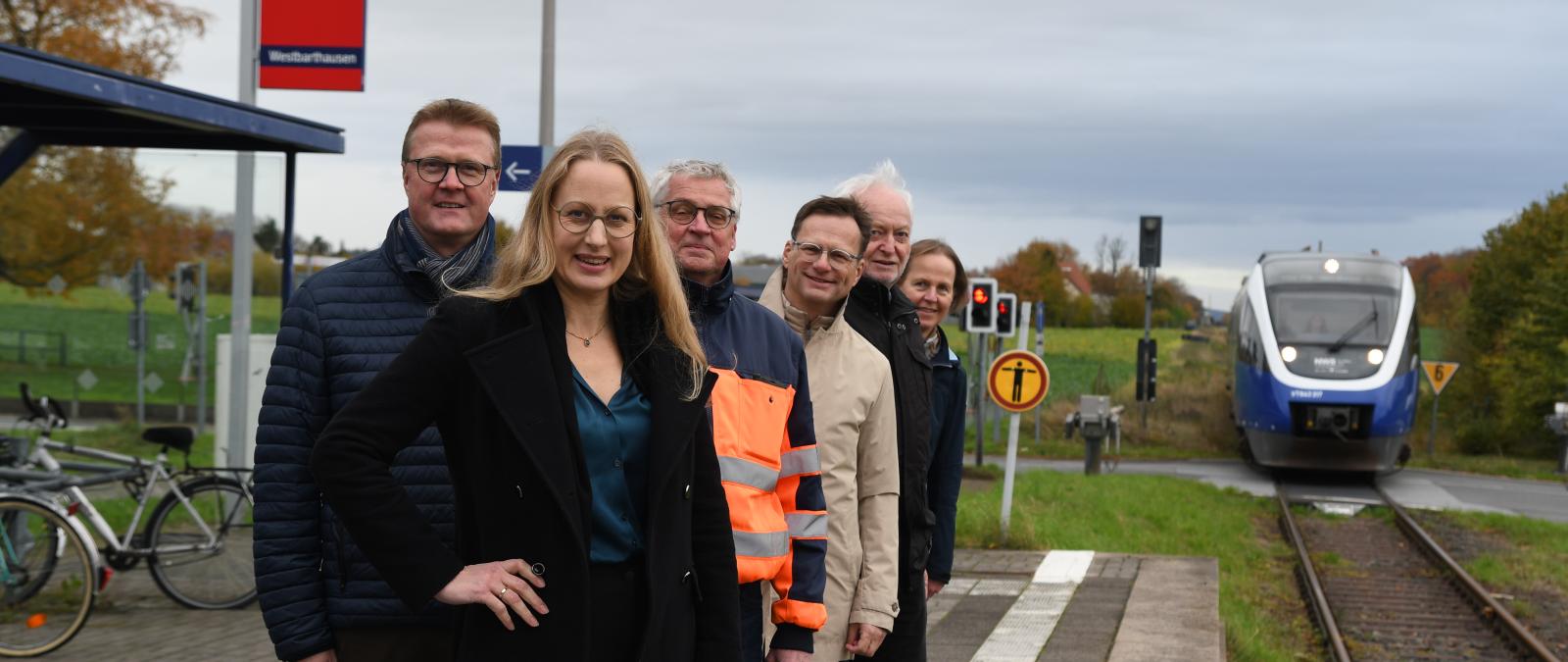 Eine Personengruppe steht auf einem Bahnsteig. Im Hintergrund nähert sich ein Zug.