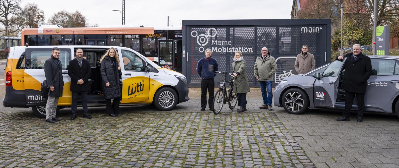 Eine Personengruppe steht vor einem Kleinbus und einem PKW.