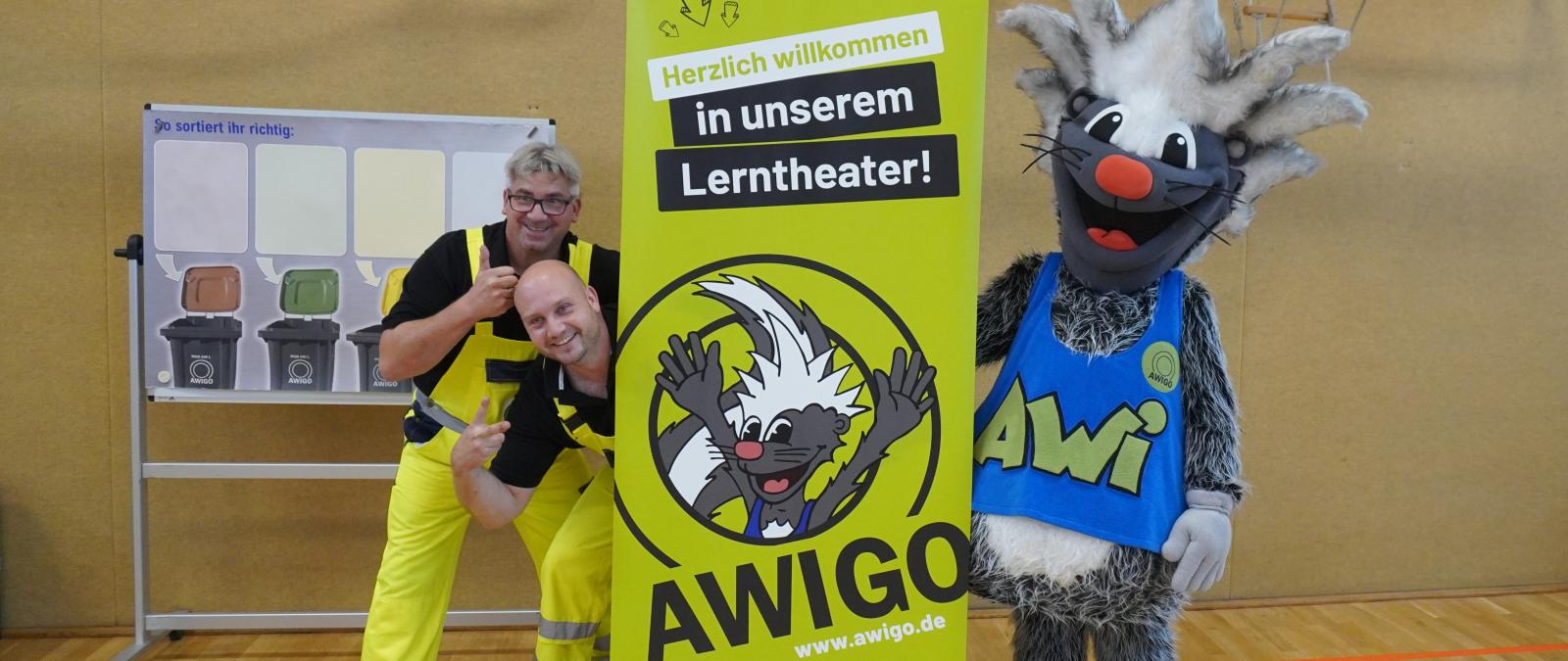 Zwei Personen und ein Maskottchen stehen neben einem Roll Up Poster.