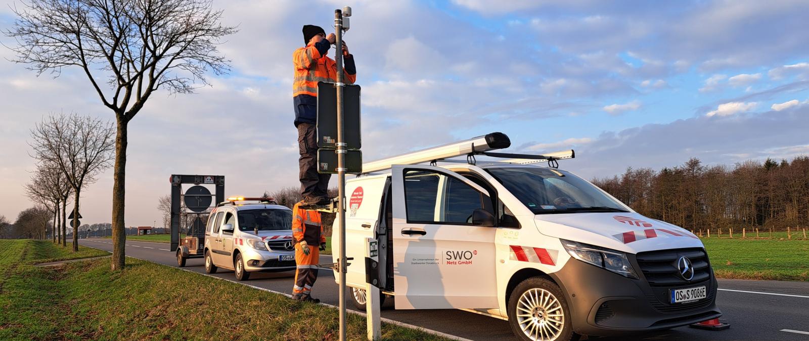 Zwei Kleinbusse parken auf einer Straße. Eine Person montiert einen Sensor an einem Mast.