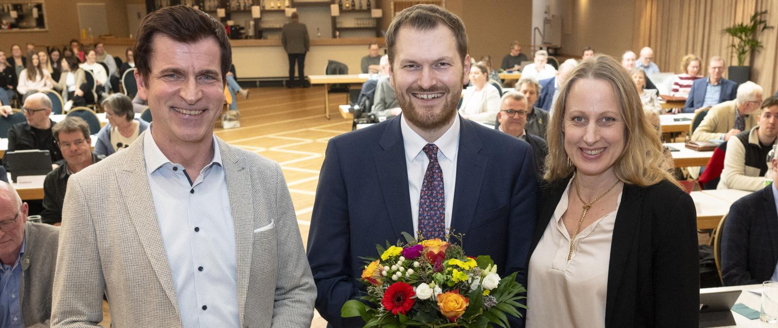 Drei Personen stehen in einem Sitzungssaal.