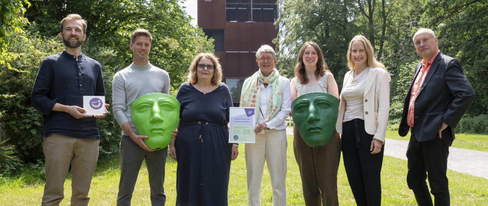 Sieben Personen mit großen römischen Masken stehen auf einer Wiese.