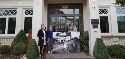 Drei Personen stehen mit einem großformatigen historischen Foto vor dem Eingang eines Hauses.