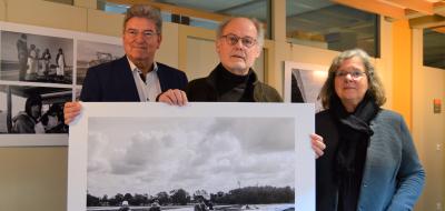 Drei Personen stehen in einem Flur und halten ein großformatiges Schwarz-weiß-Foto in den Händen.