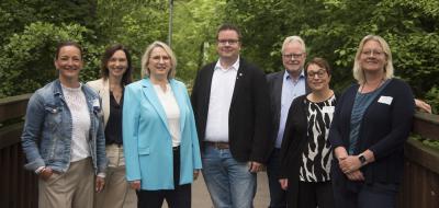 Sieben Personen stehen nebeneinander auf einem Weg mit Bäumen im Hintergrund.