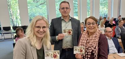 Drei Personen in einem Versammlungssaal halten Flyer in die Kamera.