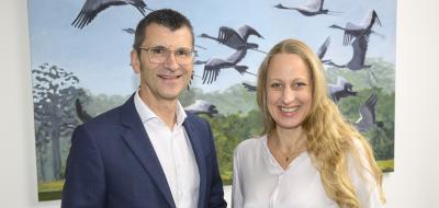 Zwei Personen stehen nebeneinander im Hintergrund ein Bild mit Vögeln.