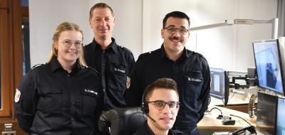 Vier Personen in Uniform stehen und sitzen in einer Rettungsleitstelle.