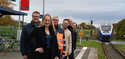 Eine Personengruppe steht auf einem Bahnsteig. Im Hintergrund nähert sich ein Zug.