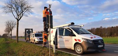 Zwei Kleinbusse parken auf einer Straße. Eine Person montiert einen Sensor an einem Mast.