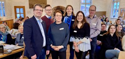 Sechs Personen stehen in einem Vortragsraum zwischen Stuhlreihen